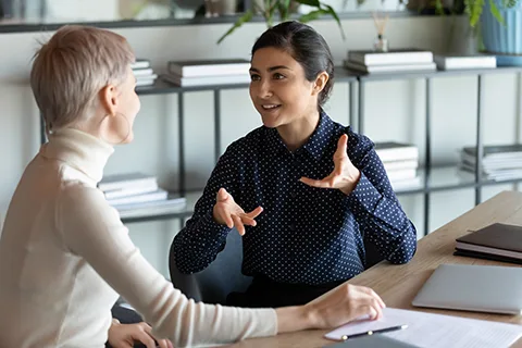 Indian female employee talking with Caucasian mate seated at workplace desk expresses her opinion on current issue